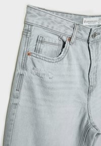 Svetlo sivi traper hlače z razfrakanim roba, obrabljenim detajlom na sprednjem žepu in bakrenimi dodatki. Ima klasičen dizajn s petimi žepi.