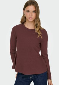 Weinrotes, geripptes Langarmoberteil mit gezacktem Ausschnitt und ausgestelltem Saum, kombiniert mit dunkelblauer Denim. Glatte Texturdetails sichtbar.