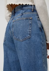 Siniset farkut korkealla vyötäröllä, joissa on kaksi takataskua, kontrastiväriset ompeleet ja lievää kulumista kankaan pinnassa.