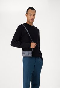 Sac en bandoulière gris avec une finition texturée et un rabat, porté sur un pull noir et un pantalon bleu clair. Design simple et rectangulaire.