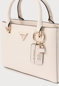 Beež tekstuuriga käekott musta ääristuse, kuldsete detailide, Guess'i logoga ja sildiga "1981." Varustatud kahe lühikese sangaga.