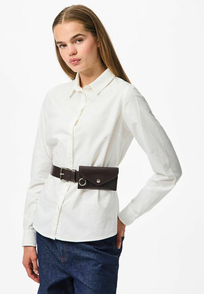 Balta pogu krekls no kokvilnas, apvienots ar brūnu ādas jostu, kurai ir kabata un apaļa metāla akcenta detaļa.