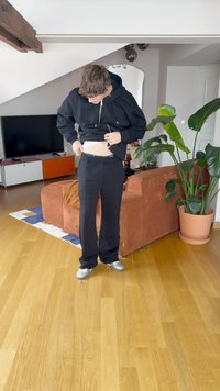Calças de treino pretas com uma cintura elástica, combinadas com um hoodie preto. O cenário inclui uma planta e móveis de cor laranja.