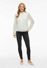 Blouse blanche avec un motif texturé en zigzag, col montant et manches longues transparentes. Associée à un jean skinny noir et des chaussures à talons légers.