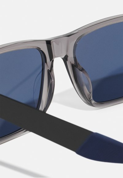 Solbriller med en gennemsigtig grå ramme, mørkeblå linser og lige sorte arme; har strukturerede marineblå accenter på tempelenderne.