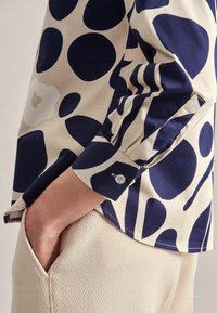 Cotton shirt featuring a cream background with large navy blue abstract dots, long sleeves, and a buttoned cuff. Textured beige pants in pocket.