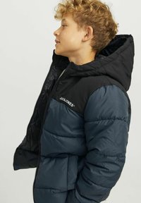 Piepildīta jaka ar kapuci, kurai ir krāsu bloku dizains melnā un tumši zilā krāsā, kā arī horizontāla quilted struktūra. Logo uz pleca.