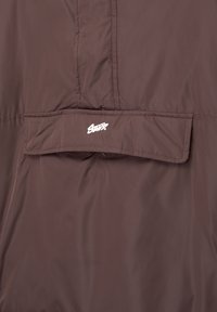 Brun vadderad jacka med hög krage, slät textur och en vit broderad logotyp på baksidan. Enkel design med minimala accenter.