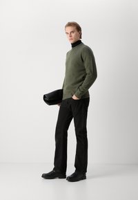 Donkergroene wollen trui met een hoge hals, gecombineerd met zwarte denim jeans en zwarte instapschoenen, terwijl ze een zwart clutch-tasje vasthoudt.