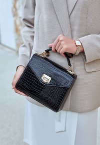 Schwarze Handtasche mit Krokodilmuster, strukturierter Form, goldenen Beschlägen und einem oberen Griff. Getragen mit einem karierten Blazer und einem weißen Oberteil.