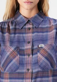 Woman wearing a blue and purple plaid button-up shirt with a collar and two chest pockets, one labeled Wrangler.
