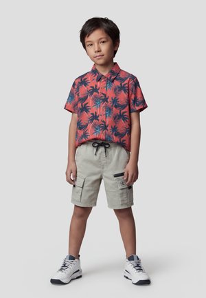 Niño de pie con las manos relajadas, vistiendo una camiseta roja de manga corta con palmeras azules, pantalones cortos cargo beige y zapatillas blancas.