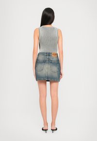 Mulher com cabelo longo e escuro, vestindo uma blusa cinza sem mangas com relevos, uma saia mini de denim desbotada e mules pretos de salto, de pé de costas.