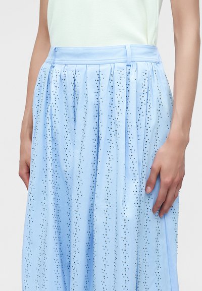 Jupe plissée bleu clair avec de petits points sombres éparpillés, portée avec un haut blanc, montrée de la taille jusqu'à la mi-cuisse avec une main reposant sur le côté.