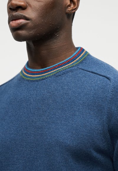 Gros plan sur un homme portant un pull bleu avec un col côtelé orné de rayures rouges, orange, jaunes et vertes.