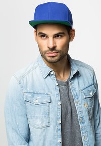 Blå baseballcap med flad skygge, båret med en lys denimskjorte over en grå T-shirt. Skjorten har knaplukning på brystlommen.