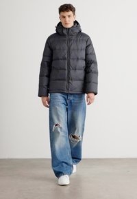 Donzen jas in donkergrijs met een capuchon, met een doorgestikt ontwerp. Gecombineerd met verwaste lichtblauwe jeans en witte sneakers.