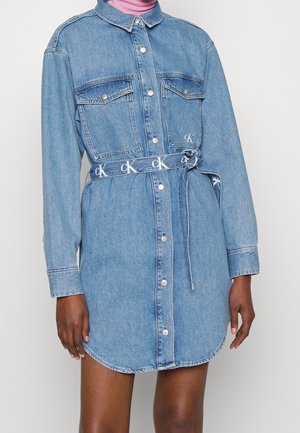 Robe-chemise en denim avec boutons sur le devant, poches poitrine et ceinture nouée avec logos CK, portée sur un col roulé rose.