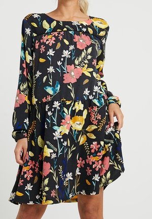 Femme portant une robe noire à manches longues avec un motif floral coloré, tenant un côté de la jupe légèrement relevé.