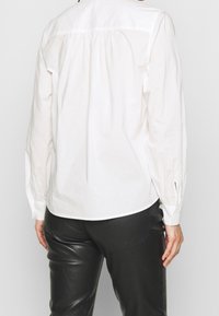 Osoba ubrana w luźną białą koszulę z długim rękawem i dopasowane czarne skórzane spodnie, stojąca tyłem na tle jasnym.