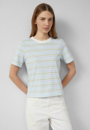 Jonge vrouw met lang lichtbruin haar, gekleed in een lichtblauw met gele gestreepte shirt met korte mouwen en een witte broek, staand tegen een grijze muur.