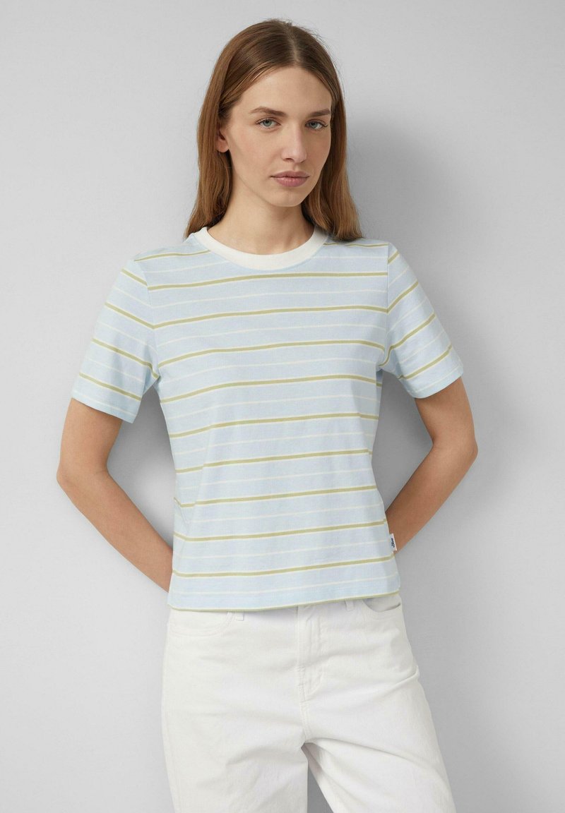 Jonge vrouw met lang lichtbruin haar, gekleed in een lichtblauw met gele gestreepte shirt met korte mouwen en een witte broek, staand tegen een grijze muur.