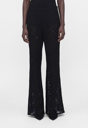 Sorte højtaljede hæklet blondebukser med blomsterprint, vide ben, båret med sorte spidse sko og en sort top.