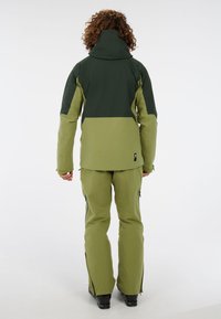 Zelené a tmavozelené dvojdielne vonkajšie oblečenie s vysokým golierom, zipsami na nohaviciach a voľným strihom. Vyrobené z vodoodpudivého materiálu.