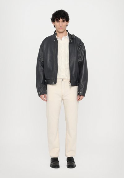 Blouson en cuir noir, pull en tricot beige et pantalon beige clair. Chaussures noires, debout sur un fond uni.