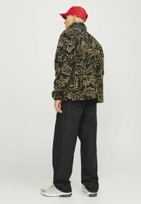 Chaqueta de forro polar con patrón negro y marrón, de corte relajado, combinada con jeans negros holgados y zapatillas blancas. Una gorra roja completa el look.