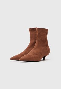 Stivali alla caviglia in suede marrone con design a punta, tacco kitten basso e superiore senza cuciture, caratterizzati da una texture liscia e senza hardware visibile.
