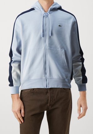 Uomo che indossa una felpa con cappuccio azzurra con zip, con strisce blu scuro e bianche sulle maniche, e pantaloni marroni, in piedi contro uno sfondo neutro.