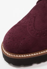 Gros plan sur le bout d'une chaussure en daim bordeaux avec des perforations style brogue, une couture détaillée, une semelle intermédiaire marron et une semelle extérieure noire texturée.