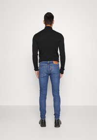 Un maglione nero a coste abbinato a jeans di denim blu. Caratteristiche distintive includono una toppa rossa con logo e stivali scuri e robusti.