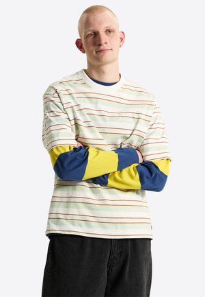 Giovane uomo con capelli corti che indossa una camicia a maniche corte a righe sopra una camicia gialla e blu a maniche lunghe, in piedi con le braccia incrociate.