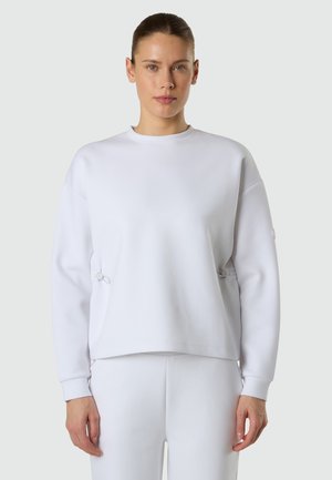Femme aux cheveux tirés en arrière, portant un sweat-shirt blanc à manches longues et un pantalon blanc assorti, debout devant un fond clair uni.