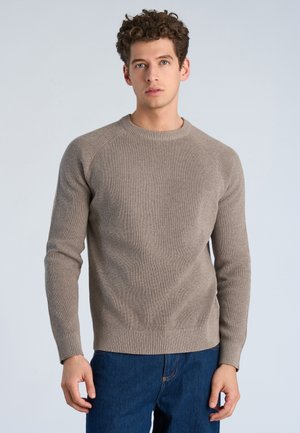 Giovane uomo con capelli ricci che indossa un maglione beige a coste e jeans blu, in piedi davanti a uno sfondo semplice grigio chiaro.