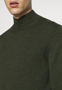 Gros plan d'une personne portant un pull à col roulé côtelé vert olive avec un petit logo brodé sur la poitrine.