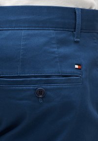 Zbliżenie na tylną kieszeń niebieskich spodni z zapięciem na guzik oraz małe prostokątne logo marki w kolorach czerwonym, białym i czarnym nad kieszenią.