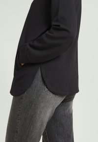 Maglia nera a maniche lunghe con orlo curvo, caratterizzata da fessure laterali. Abbinata a jeans in denim grigi chiari, con dettaglio della tasca in evidenza.