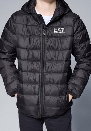 Personne portant une veste matelassée noire EA7 Emporio Armani avec capuche, fermeture éclair fermée, mains dans les poches, debout devant un fond uni.
