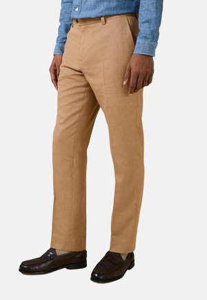 Man draagt lichtbruine nette broek, donkerbruine loafers en een blauw overhemd met knopen, staand tegen een witte achtergrond.