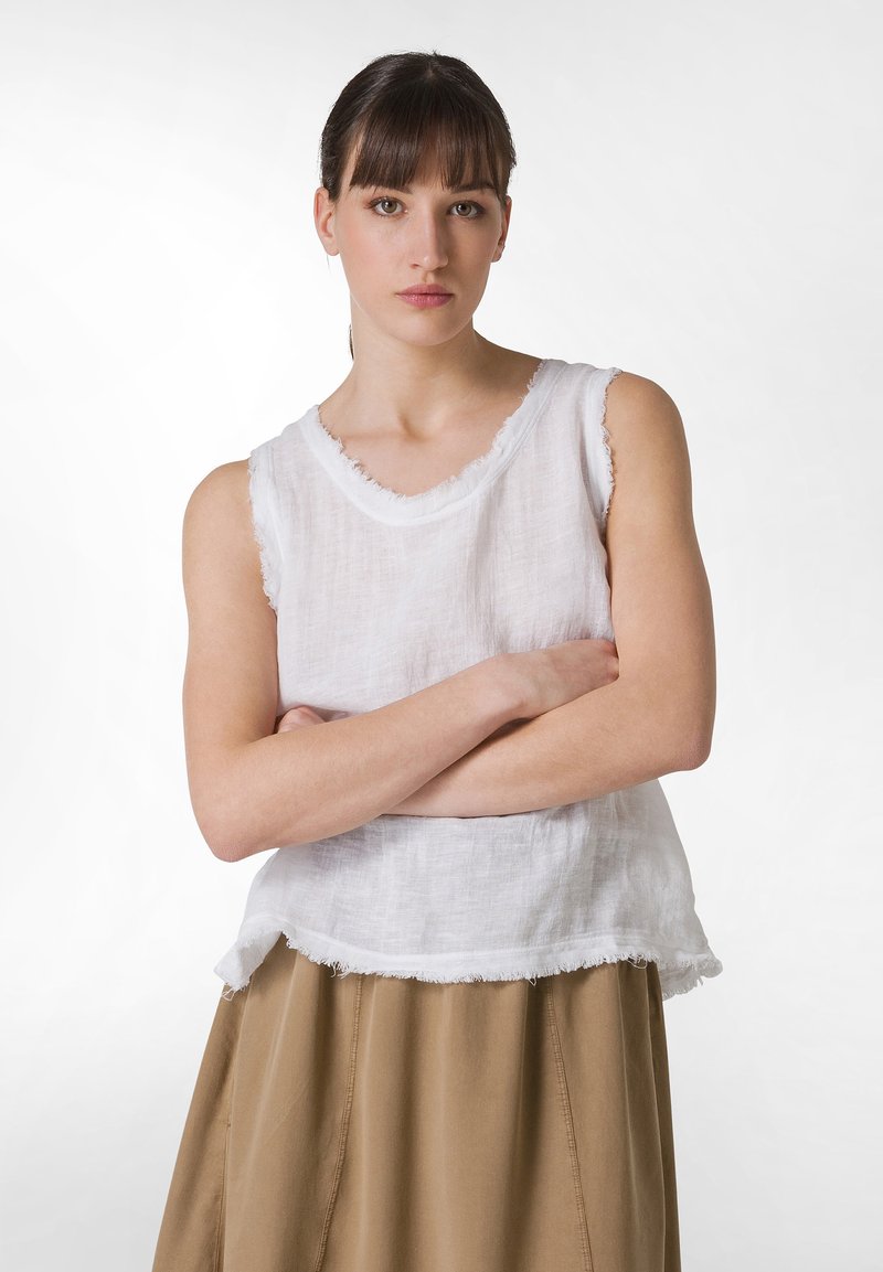 Witte linnen mouwloze top met een ruwe zoom. Model staat met gekruiste armen en draagt een tan rok met een uitlopende silhouette. Eenvoudig ontwerp.