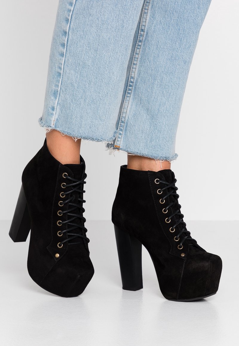 Bottines montantes à lacets en daim noir à plateforme portées avec un jean bleu clair raccourci à ourlet effiloché, sur un sol blanc.
