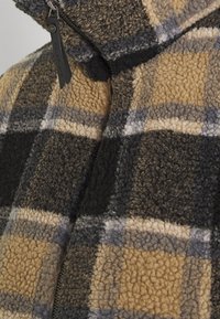 Brown and gray checkered fleece jacket with a high collar, featuring a zipper and textured material. Accents of black and cream visible.