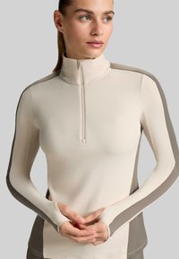 Hellbeiges Langarmoberteil mit halbem Reißverschluss, dunkelbeigen Seitenpaneelen und Daumenlöchern, aus glattem, dehnbarem Stoff gefertigt.