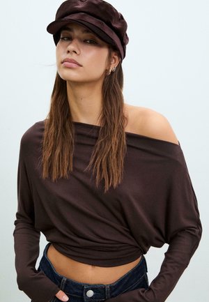 Brązowa bluzka typu off-the-shoulder z długim rękawem o miękkiej, zwisającej fakturze, zestawiona z ciemnobrązową czapką i ciemnymi dżinsami z zapięciem na guzik.