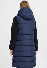 Mørkeblå puffervest med en fitted design, som har hette og svarte, fleece-lignende ermer. Bak viser den quiltede seksjoner og kontrasterende teksturer.