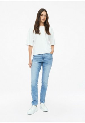 Junge Frau mit langen dunklen Haaren, die ein weißes übergroßes Kurzarmshirt, hellblaue Jeans und weiße Sneaker trägt und mit der Hand in der Tasche steht.