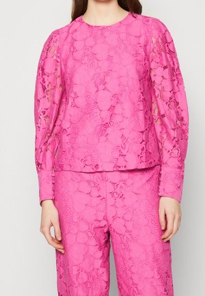 Mujer con top de encaje rosa brillante y pantalones a juego con patrones florales recortados y mangas largas abullonadas.